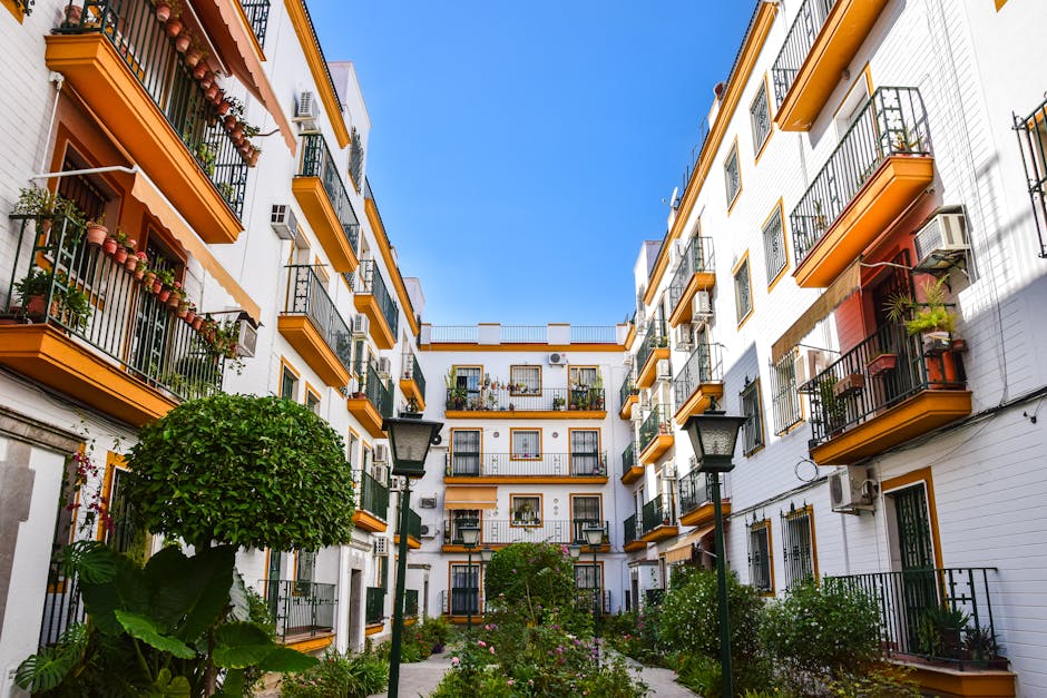 A view of a small, landscaped courtyard within a multi-story residential building complex, featuring a central paved pathway flanked by neatly maintained garden beds filled with lush green shrubs, flowering plants, and small trees. The buildings surrounding the courtyard are painted white with accents of yellow-orange on the balconies’ trims and window frames, some with potted plants and outdoor furniture. Several black wrought-iron balcony railings can be seen, each adorned with potted flowers and greenery. Traditional street lamps line the pathway, providing lighting for evening visits. The sky above is clear and bright blue, indicating a sunny day, and the overall scene showcases urban garden maintenance and landscaping work often performed by Gardeners Pimlico for balcony and courtyard gardens.