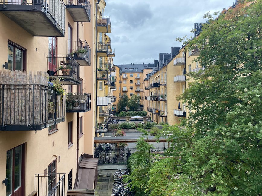 A view of a small, landscaped courtyard within a multi-story residential building complex, featuring a central paved pathway flanked by neatly maintained garden beds filled with lush green shrubs, flowering plants, and small trees. The buildings surrounding the courtyard are painted white with accents of yellow-orange on the balconies’ trims and window frames, some with potted plants and outdoor furniture. Several black wrought-iron balcony railings can be seen, each adorned with potted flowers and greenery. Traditional street lamps line the pathway, providing lighting for evening visits. The sky above is clear and bright blue, indicating a sunny day, and the overall scene showcases urban garden maintenance and landscaping work often performed by Gardeners Pimlico for balcony and courtyard gardens.