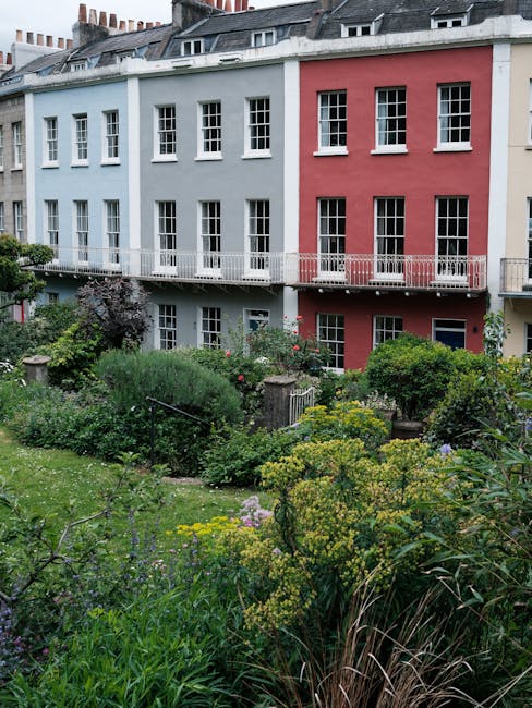 A small, well-maintained garden featuring a paved patio area with reddish-brown stone tiles, bordered by neatly trimmed rectangular and square hedges filled with low-growing green foliage. In the center, a decorative wrought-iron garden bench with intricate scrollwork and a dark finish faces the patio, positioned in front of a white building with a centered window framed by white shutters and a small black wrought-iron balcony. To the left, a large leafy tree with a textured trunk extends its branches over the garden, creating shade and adding to the lush greenery. On the right, a bushy shrub with dense foliage partially obscures wooden steps ascending to a side entrance, while various plants, including ferns and flowering perennials, fill the garden beds along the wall and steps. The scene is lit softly, suggesting late afternoon or early evening, with natural light filtering through the leaves, emphasizing the textures of the soil, plants, and aged stone paving. Gardeners Pimlico, a professional gardening service, maintains this outdoor space, which exemplifies tidy lawn care, hedge trimming, and garden maintenance typical of landscaped residential gardens.